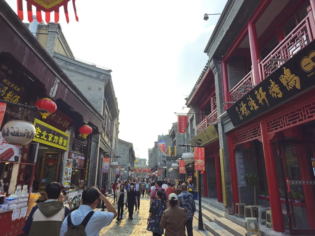 Lantern-lit hutong alley in a Beijing old neighbourhood with people walking past low-rise buildings.